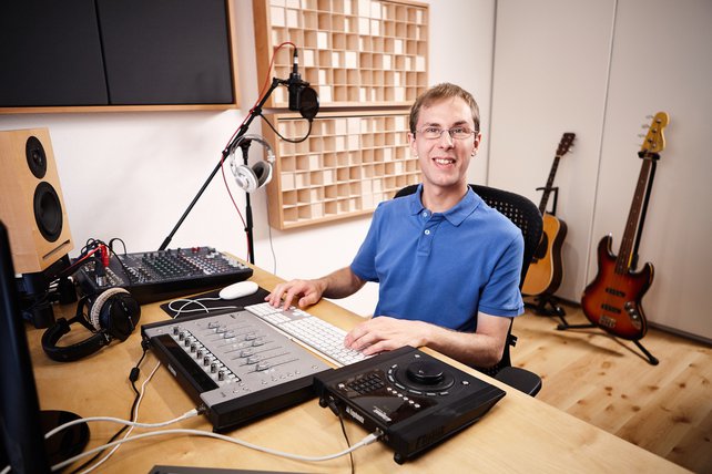 Florian Schmitz sitzt am Studio-Tisch und lächelt in die Kamera. Im Vordergrund ist die Studio-Technik zu sehen. An der Wand sind Akustik-Module montiert und im Hintergrund stehen ein E-Bass und eine Akustik-Gitarre.