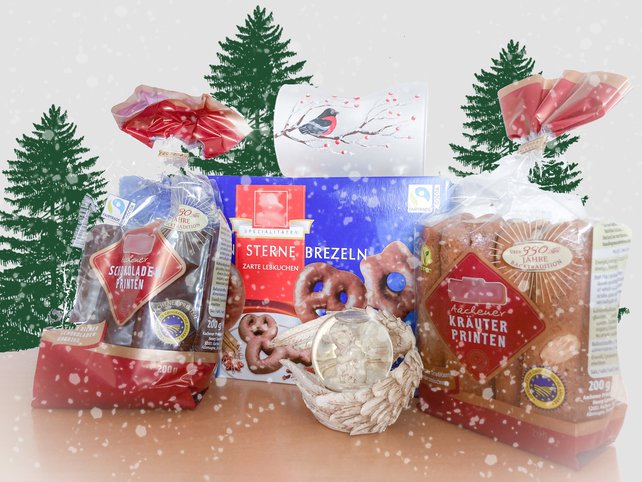 Zwei Packungen Printen stehen auf einem Tisch angelehnt an eine Packung Lebkuchen. Es ist weihnachtlich dekoriert. Im Hintergrund sind drei gemalte Tannenbäume zu sehen und es schneit. Das Ganze ist absichtlich kitschig gestaltet.